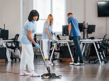 Woman uses vacuum cleaner. Group of workers clean modern office together at daytime.