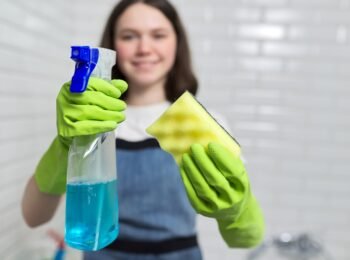 Portrait of girl doing cleaning in bathroom. Teenager in apron gloves with detergent and rag sponge smiles looking at camera. Housekeeping, cleanliness at home, service, young people concept