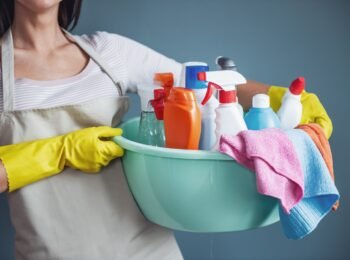Cropped image of beautiful woman holding a basin with detergents and rags, on gray background