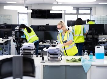 Caucasian woman and colleagues wearing hi vis vests, gloves, safety glasses and face masks sanitizing an office using disinfectant. Hygiene in workplace during Coronavirus Covid 19 pandemic.