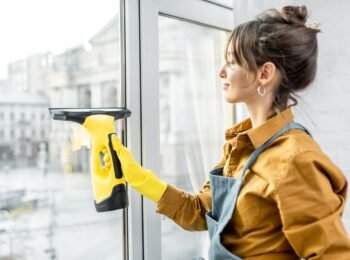 Beautiful young housewife in working clothes washing windows by professional tools at home